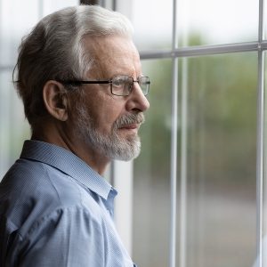 Older man looking pensively out a window implying feeling lost or depressed. He's thinking about starting therapy.