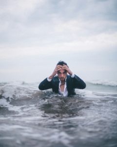 Man in a suite but in the ocean with water up to his torso. Hand on head in frustration and he's thinking he needs a therapist
