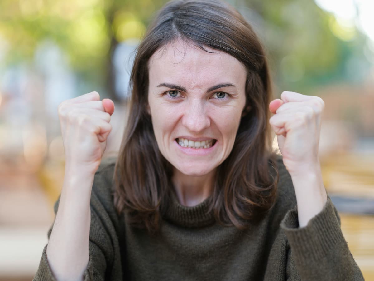 A close-up of a woman with a frustrated facial expression, gritting her teeth and holding up two clenched fists toward the camera.