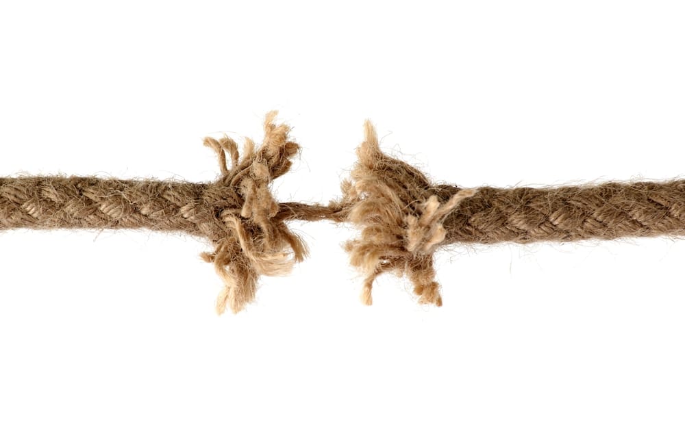 A close-up photograph of a brown natural fiber rope that is severely unraveled, frayed, and nearly broken in the middle, held together by only a thin internal cord, symbolizing the extreme strain and breaking point of burnout.