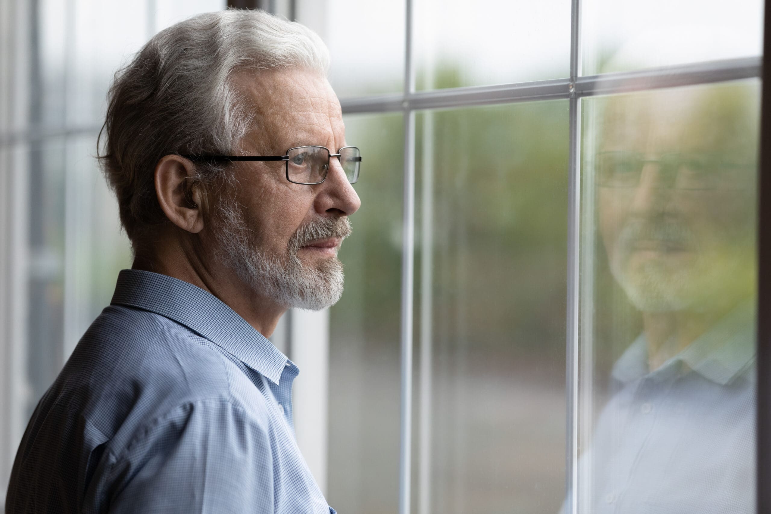 An older man with glasses looking thoughtfully out a window.
